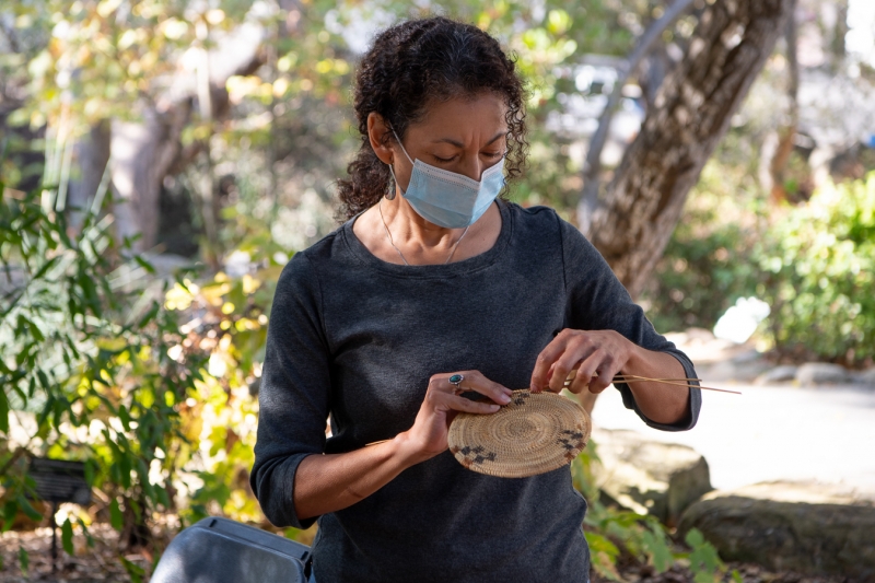 A woman with dark, curly hair works on the base of a basket. The design looks like checks, they are the beginning of spirals.