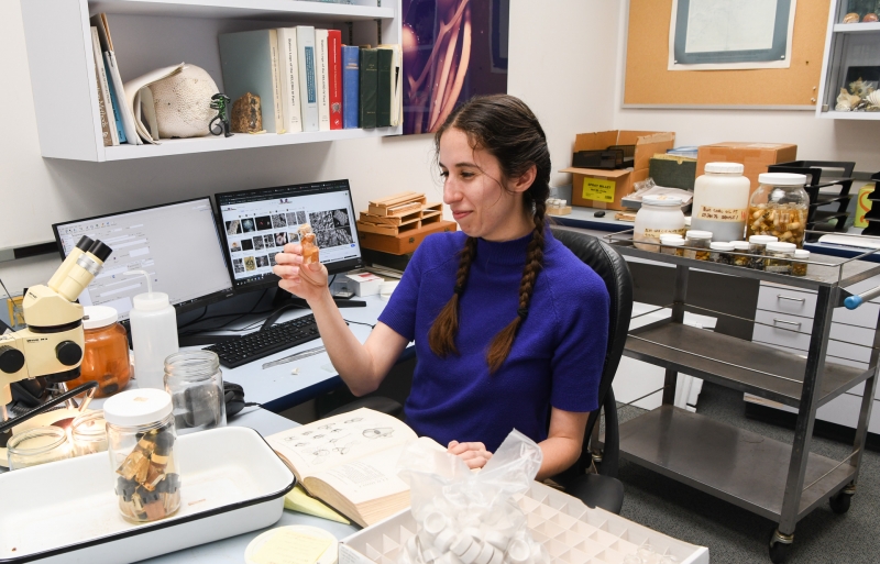 A smiling woman looks at a tiny vial, surrounded by books, computer, microscope, and other scientific paraphernalia