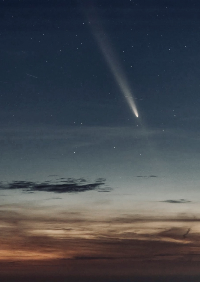 The white tail of a comet streams across a sky with clouds and a final glow of sunset